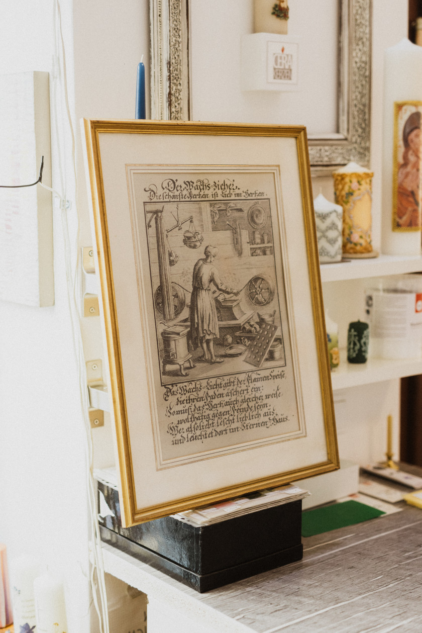 Auf diesem Foto steht ein Bild im Mittelpunkt, das goldgerahmt ist. Auf dem Bild ist das Wachsziehen zu sehen und dann etwas in alter Schrift geschrieben. Das Foto ist Schwarz-Weiß und sieht sehr alt aus. Es steht auf einer schwarzen Box. Im Hintergrund sind Regale mit Kerzen zu sehen.