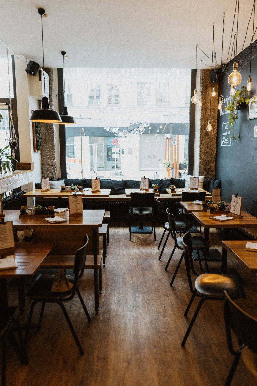 Auf diesem Foto sieht man einen Blick in den Laden. Links und rechts sind Stühle mit Tischen zu sehen. Ein kleiner Gang dazwischen führt zum Schaufenster des Restaurants. Vor dem Schaufenster sind ebenfalls Tische und eine Bank zu sehen. Über den Tischen hängen Lampen von der Decke. Die Tische und Stühle sind aus Holz. Links im Hintergrund ist ganz wenig noch die Eingangstüre zu erkennen. Am linken Rand des Fotos sind Fliesen und eine halbhohe Mauer zu erkennen. Rechts im Bild am Rand ist eine dunkle Wand zu erkennen, an der etwas angebracht wurde. Im Hintergrund sieht man durch das Schaufenster nach draußen.