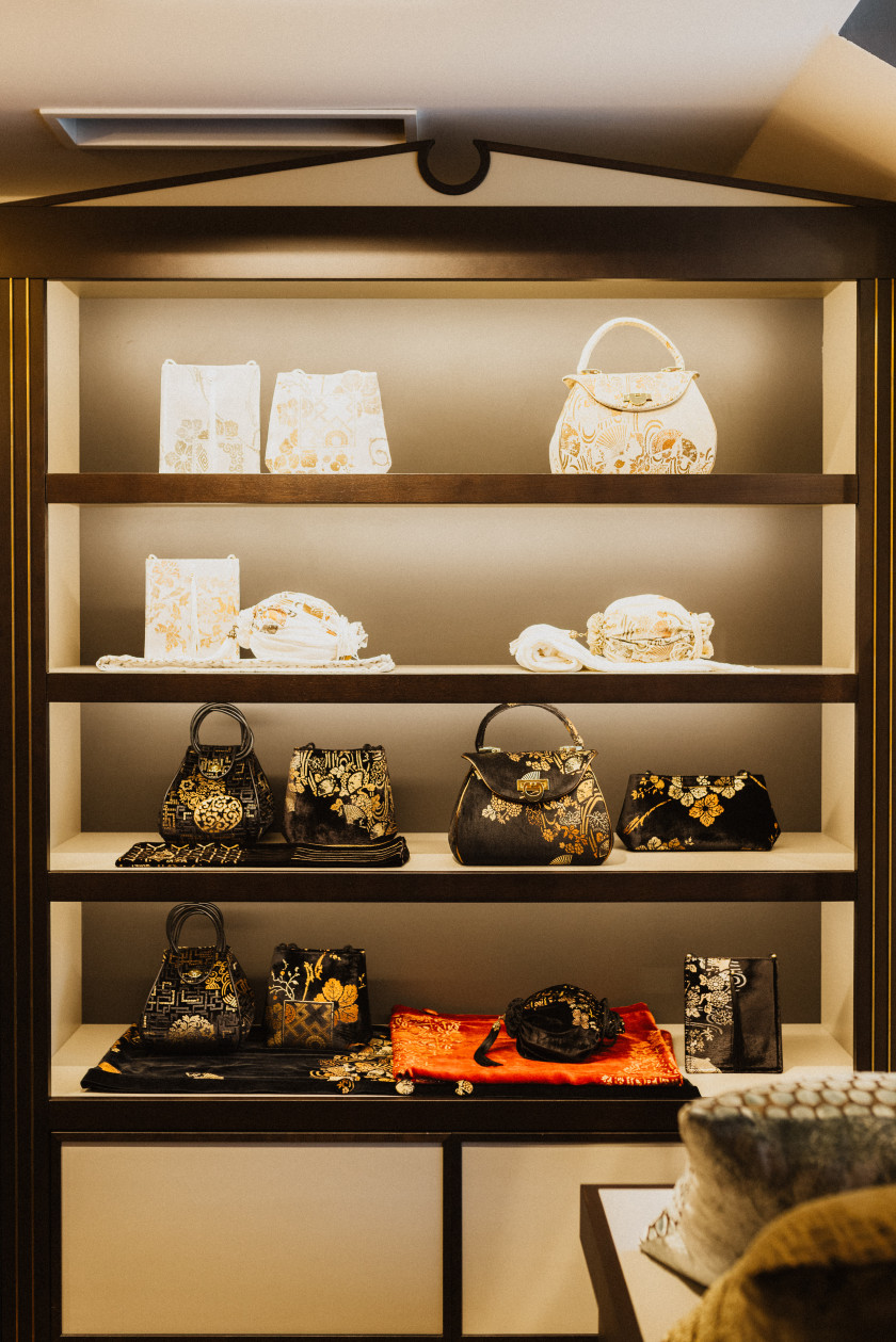 Auf diesem Bild sind die Taschen im Vordergrund. Diese stehen aufgereiht in einem Regal und werden beleuchtet. Die Taschen sind aus Samt und mit edlen Mustern.