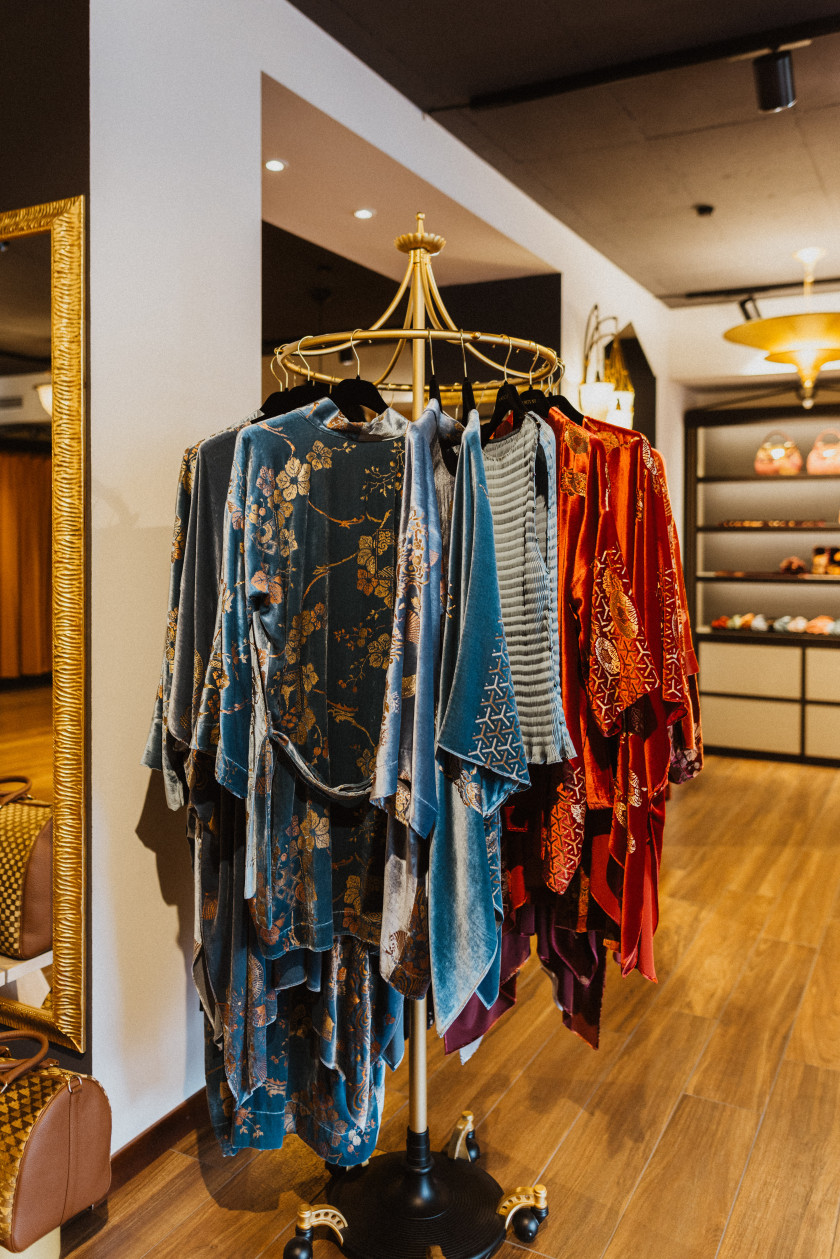 Auf diesem Foto sieht man im Vordergrund einen Kleiderständer, an dem verschiedene Kimonos an Kleiderbügeln hängen. Der Kleiderständer ist Gold. Die Kimonos sind in allen verschiedenen Farben und Mustern. Die Farben Blau und Rot stehen im Fokus. Im Hintergrund ist verschwommen ein Regal zu sehen, an der Decke eine Lampe. Links an der Seite im Vordergrund sind ein abgeschnittener Spiegel und eine Tasche.