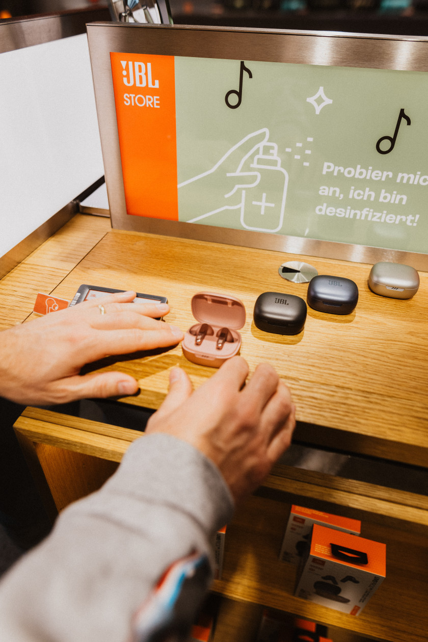 Auf diesem Foto sieht man zwei Arme, die von unten ins Bild ragen und man blickt auf eine Holzablage eines Regals. Darauf liegen In-Ear Kopfhörer in verschiedenen Farben. Drei sind in geschlossenen Cases. Eines ist offen und man sieht die Kopfhörer darin liegen. Die Hände berühren das Case von außen. An dem Regal ist ein Schild befestigt auf dem steht „Probier mich an, ich bin desinfiziert!“ und ein Symbol mit Hand und Spray sowie Noten.