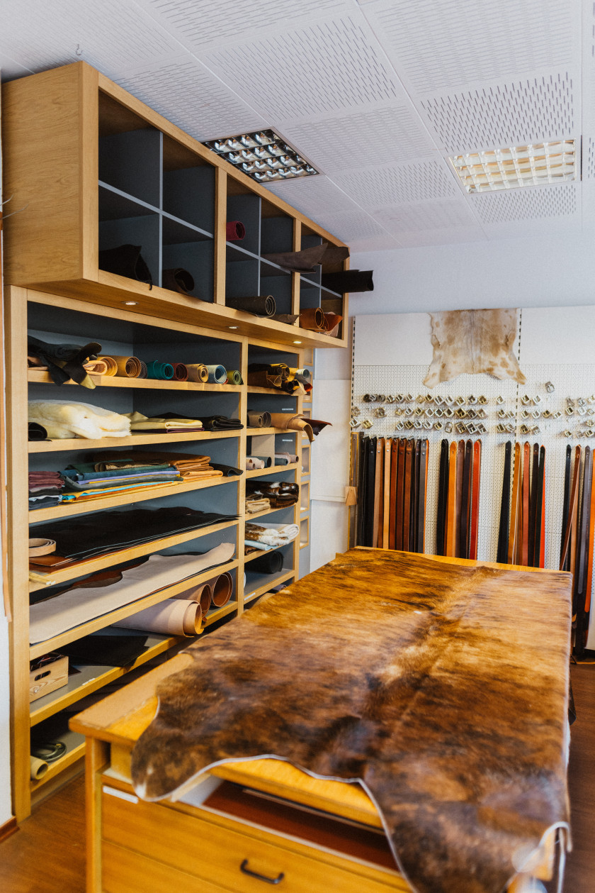 Auf diesem Bild sieht man einen Blick in den Laden. Im Zentrum steht ein Tisch mit Schubladen, auf dem ein Tierfell liegt. Im Hintergrund sind Gürtel und Gürtelschnallen an der Wand aufgehängt, darüber noch ein Tierfell. Links auf dem Foto ist ein deckenhohes Regal zu sehen. In dem Regal befinden sich verschiedene Leder aufgerollt oder liegend übereinander.