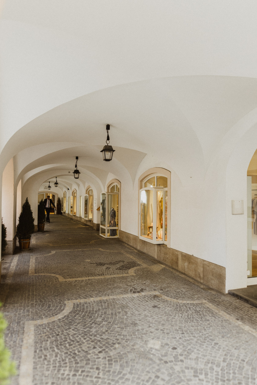 Hier sieht man den Laden von außen, der unter den Bögen des Hauses liegt. Rechts an den Wänden sieht man die Schaufenster. Davor sind Pflastersteine und ein weiter Gang zu sehen. An der Decke hängen Lampen. Im Mittelpunkt des Bildes stehen der Blick durch die Bögen und die seitliche Ansicht des Geschäftes unter den Bögen.