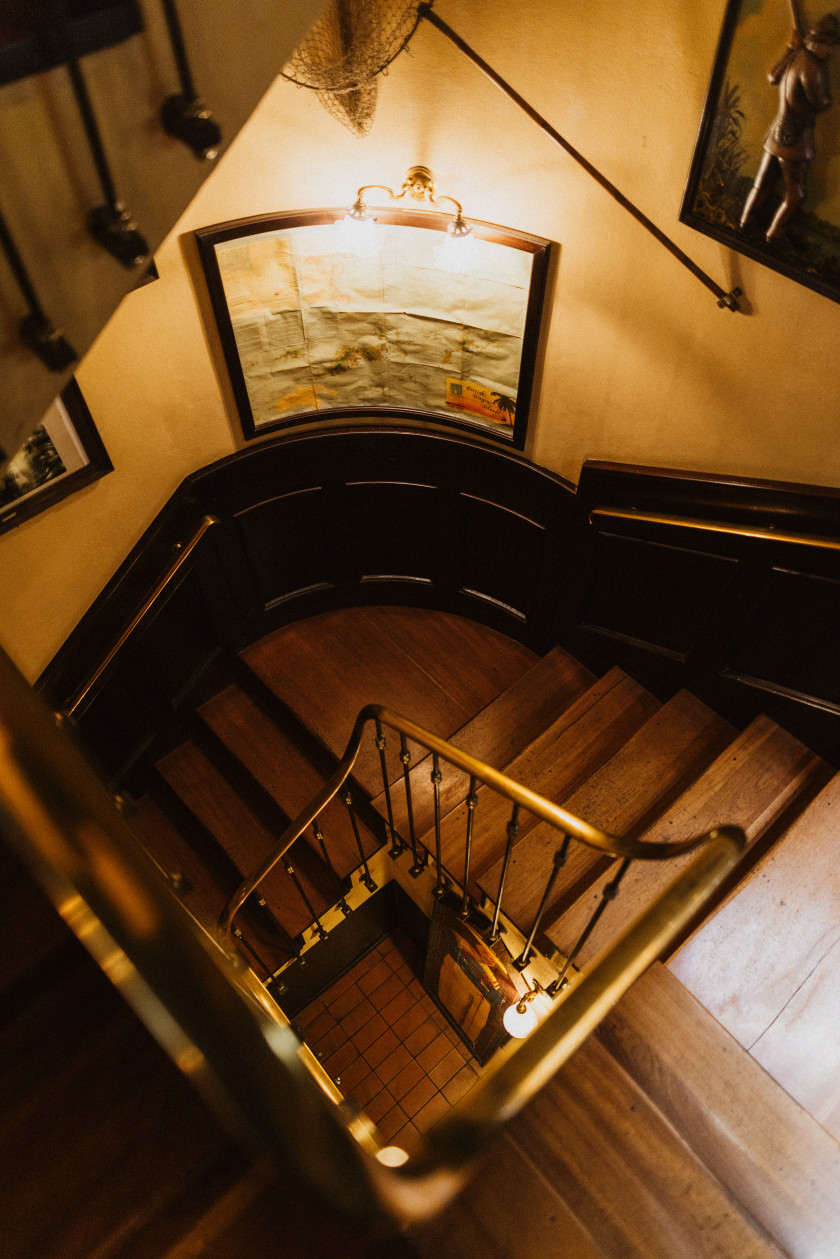 Auf diesem Bild sieht man eine Treppe aus dunklem Holz, die hinunterführt. Das Geländer der Treppe ist Gold. Über den Treppenstufen an der Wand ist eine dunkle Holzvertäfelung. Die Wand darüber ist gelb und dort ist ein Bild befestigt, das angestrahlt wird von einer Lampe, die ebenfalls an dieser Wand über dem Bild befestigt ist.