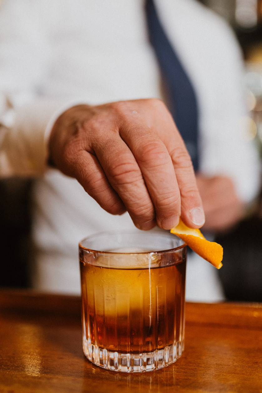 Auf diesem Foto sieht man ein Glas auf einem Bartresen stehen. Der Tresen ist aus dunklem Holz. Das Glas ist gefüllt mit einem Getränk und darin liegt ein runder, großer Eiswürfel. Das Getränk ist braun. Hinter der Bartheke ist verschwommen ein Oberkörper mit weißem Hemd und schwarzer Krawatte zu sehen. Man sieht eine Hand an dem Glas, die ein Stück einer Orangenschale hält und an den Rand des Glases reibt.