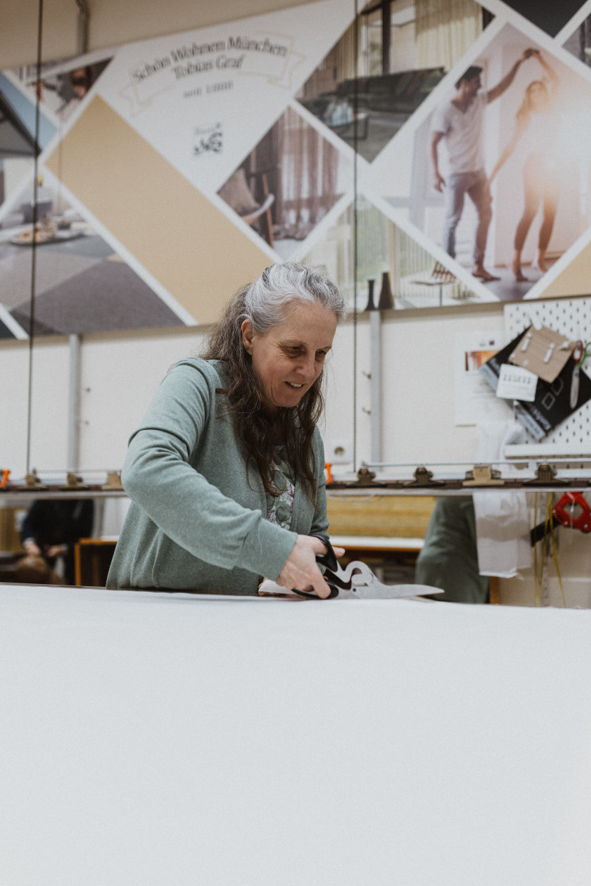 Auf diesem Foto sieht man Monika Rothammer, eine Mitarbeiterin von Schön Wohnen, im Vordergrund. Sie trägt einen grünen Cardigan und ist fokussiert auf ihre Arbeit, lächelt dabei aber leicht. Sie hält eine Schere in ihrer Hand und schneidet gerade einen weißen Stoff. Im Hintergrund ist das Nähatelier zu erkennen.