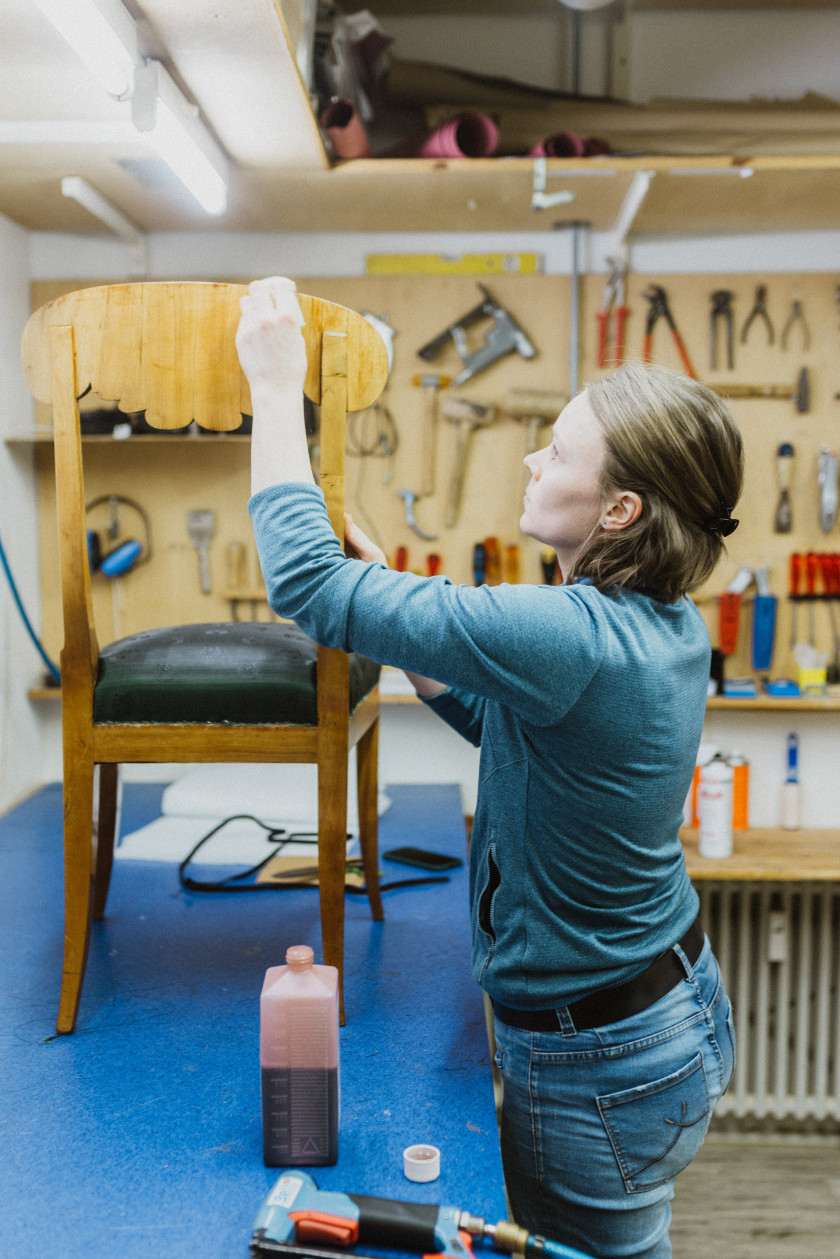 Auf diesem Foto sieht man, wie eine junge Frau in einer Werkstatt vor einer blauen Arbeitsplatte steht. Das Foto wurde mit seitlichem Blick auf die Arbeitsplatte und auf die junge Frau aufgenommen. Auf dem Tisch vor der Frau steht ein Stuhl aus Holz mit einer grünen Sitzfläche. Die junge Frau trägt eine Jeans und ein blaues Sweatshirt. Mit ihren beiden Händen berührt sie den Stuhl und es sieht aus, als ob sie diesen abschleift oder poliert. Vor ihr auf der Arbeitsfläche stehen außerdem ein Behälter mit roter Flüssigkeit und ein anderes Gerät. Die junge Frau blickt konzentriert auf ihre Arbeit und den Stuhl. Im Hintergrund sieht man eine Wand, an der verschiedene Werkzeuge wie Hammer, Zangen oder Schraubschlüssel befestigt sind. Darunter ist ein Heizkörper zu erkennen.