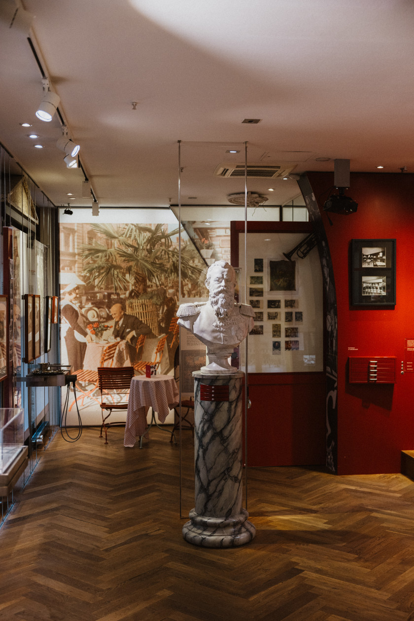 Auf diesem Foto sieht man einen Blick in das Museum des Hauses. In der Mitte des Raumes steht eine Büste eines Männerkopfes auf einer Marmorsäule. Der Boden ist aus Holz. Links an der Wand erkennt man Bilder. Rechts im Bild ist eine rote Wand mit einem Bild. Im Hintergrund an der hinteren Wand ist ein Bild über die ganze Wand einer Cafészene zu erkennen. Davor steht ein Tisch mit weißer Tischdecke und zwei roten Stühlen. An der Decke sind Strahler angebracht, die den Raum beleuchten.