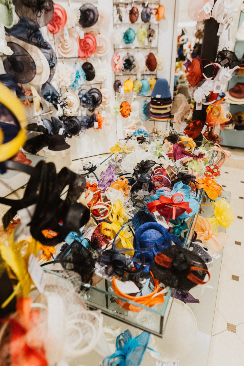 Dieses Foto wurde ebenfalls im ersten Stock des Ladens aufgenommen, was man an dem weiß gefliesten Boden mit den schwarzen Quadraten erkennt. Im Zentrum des Fotos ist ein Tisch mit einer Glasplatte, auf dem bunte, ausgefallene Hüte wie Fascinator und Schleifen zu sehen sind. Auch im Hintergrund sind Regale mit verschiedenen ausgefallenen und bunten Hüten, die dort an den Wänden hängen.