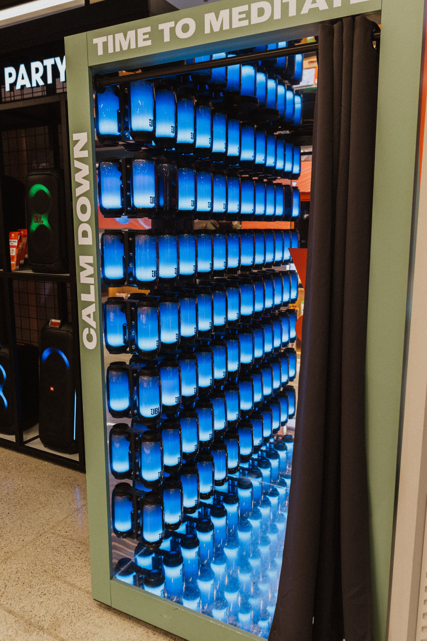 Auf diesem Foto sieht man eine Kabine mit geöffnetem Vorhang. In der Kabine sieht man an der Wand, die zu sehen ist, viele identische blau beleuchtete Musikboxen an der Wand befestigt. Diese spiegeln sich im Boden der Kabine. Außen auf der Kabine steht „CALM DOWN, TIME TO MEDI“.