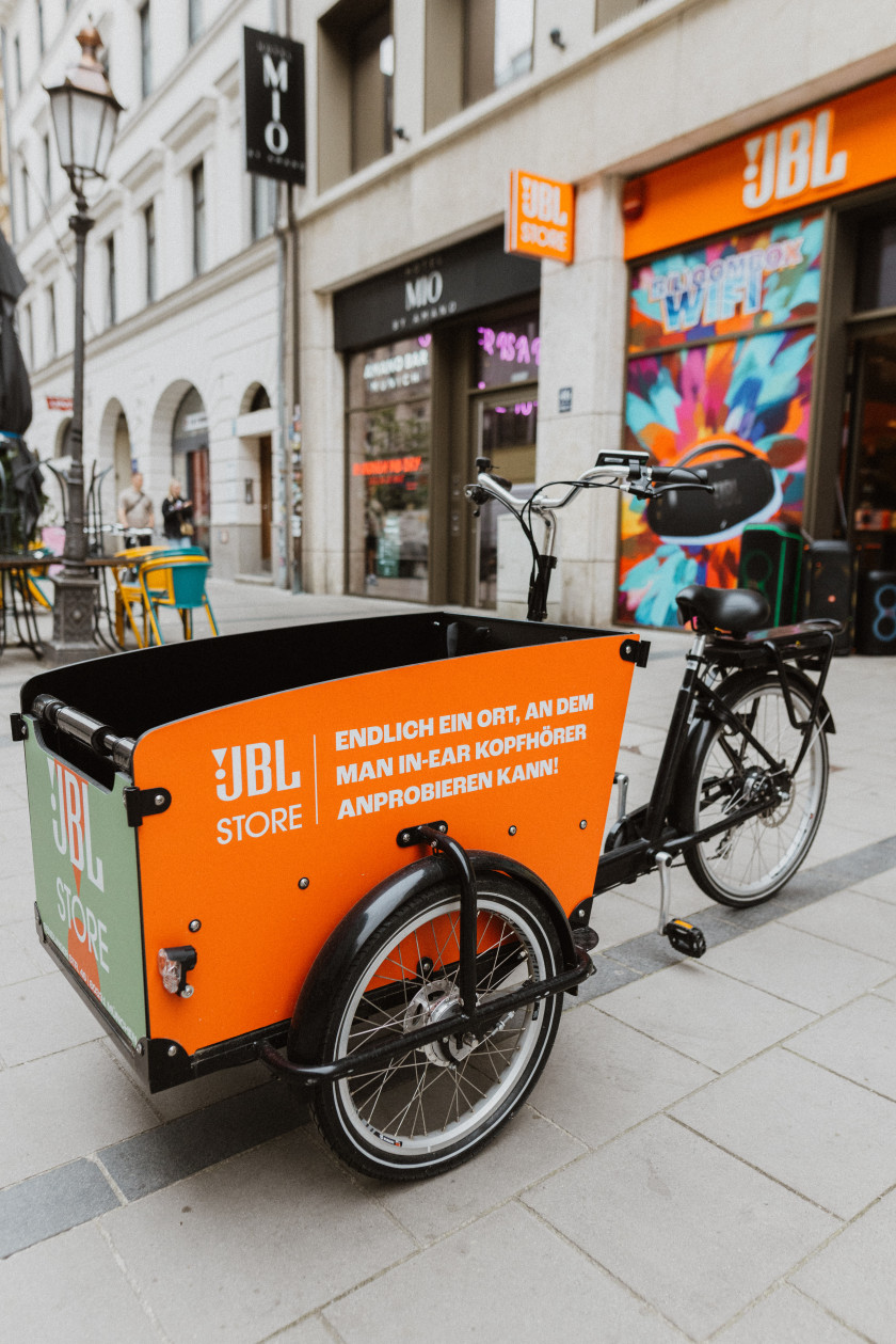 Auf diesem Foto sieht man ein Lastenfahrrad vor einem Geschäft auf der Straße stehen. Auf dem Lastentransport steht in weißer Schrift auf orangenem Hintergrund „Endlich ein Ort, an dem man In-Ear Kopfhörer anprobieren kann!“. Das Rad dahinter ist schwarz.