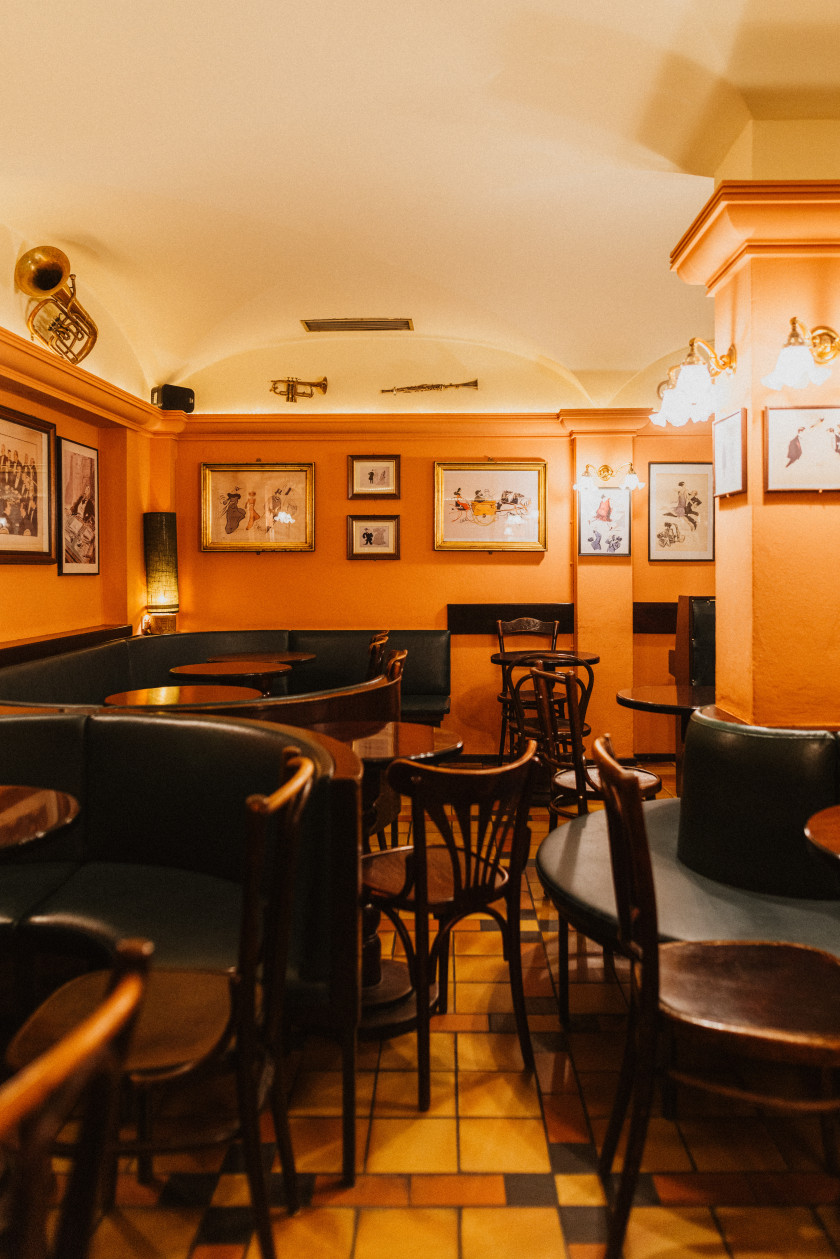 Auf diesem Foto sieht man einen Blick in die Piano-Bar im Untergeschoss. Der Raum sieht aus wie ein Gewölbe. Die Wände sind Orange gestrichen. An den Wänden hängen Bilder, Lampen und Musikinstrumente. Im Raum stehen Tische und Stühle aus dunklem Holz. Die Bänke sind an den Wänden befestigt, rund und aus dunklem Kunststoff. Der Boden ist hell gefliest.