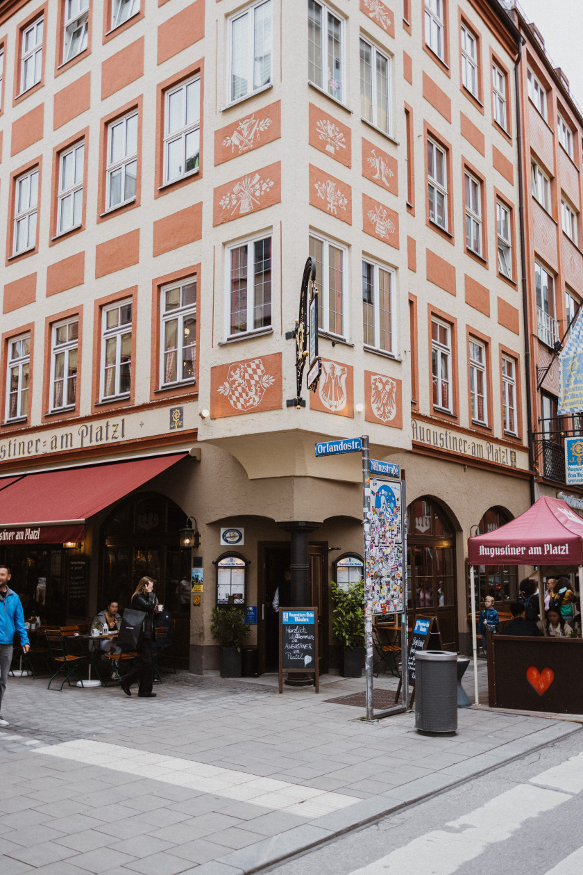 Auf diesem Foto sieht man die Außenfassade des Wirtshauses. Diese geht um eine Ecke. Das Foto wurde in Draufsicht auf diese Ecke aufgenommen. Das Haus ist orange-weiß und es sind viele Fenster zu sehen. Rechts im Bild vor dem Haus ist ein Schanigarten zu sehen mit einem roten Pavillon. Mittig sind ein Mülleimer und die Straßenschilder zu sehen. An der Ecke des Hauses ist der Eingang. Vor dem Eingang steht eine Tafel auf dem Boden. Links im Bild ist eine rote Markise ausgefahren. Darunter befinden sich Personen sowie Tische und Stühle. Unten im Bild sieht man noch einen Teil des Fußweges und der Straße. Auf dem Haus sieht man über den Fenstern links und rechts auf der Fassade den Schriftzug „Augustiner am Platzl“ sowie auf dem Erker an der Ecke verschiedene Wappen.