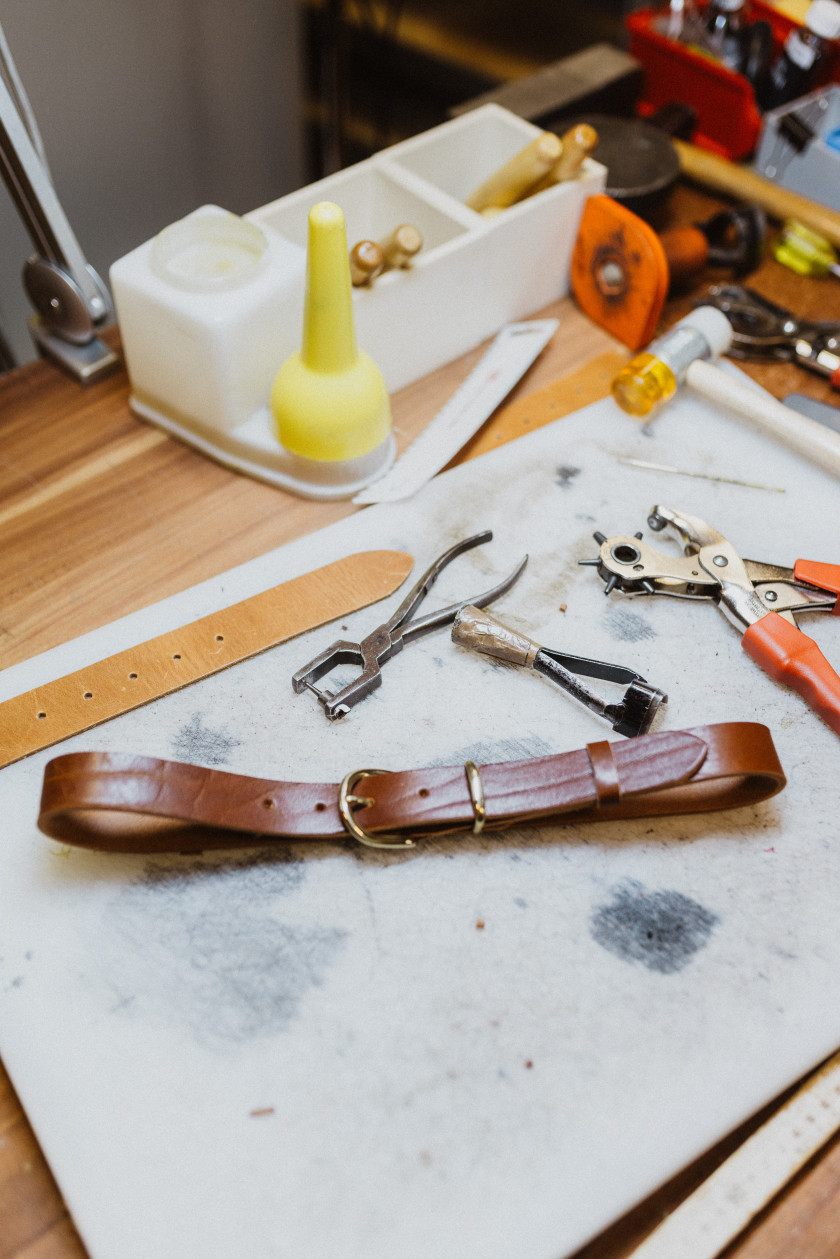 Auf diesem Foto sieht man einen Gürtel im Fokus. Dieser liegt auf einer Arbeitsfläche. Daneben sind verschiedene Geräte zur Bearbeitung von Leder. Der Gürtel ist braun und die Schnalle ist verschlossen. Neben der Arbeitsfläche liegen auf dem Schreibtisch noch verschiedene Materialien, die nicht genau zu erkennen sind.