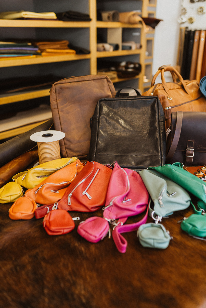Auf diesem Bild sieht man verschiedene Taschen. Vorne sind Bauchtaschen aus Leder in verschiedenen Farben zu sehen. Dahinter stehen Rucksäcke aus Leder, und ein Band aufgerollt steht daneben. Alle Taschen befinden sich auf einem braunen Fell. Im Hintergrund sind verschwommen Regale mit Leder zu erkennen.