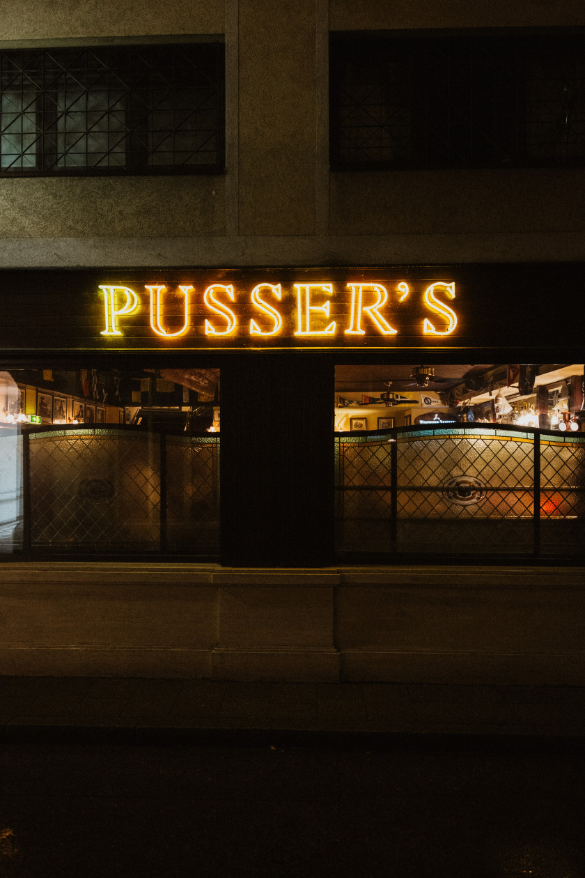 Auf diesem Foto sieht man einen Blick auf die Außenfassade der Bar bei Nacht. Es sind die Fenster und dahinter leichtes Licht zu erkennen. Über den Fenstern steht in orangenen Leuchtbuchstaben der Schriftzug „PUSSER´S“.