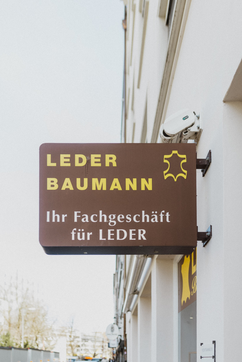 Auf diesem Bild sieht man den Aushänger von Leder Baumann an der Fassade vor dem Laden. Auf dem Aushängeschild steht „Leder Baumann – Ihr Fachgeschäft für LEDER“ in gelber und weißer Schrift auf braunem Hintergrund. Daneben ist das Logo in Gelb zu sehen, welches an ein Kuhfell erinnert. Im Hintergrund ist der Verlauf der Herzog-Wilhelm-Straße zu sehen und der Himmel.