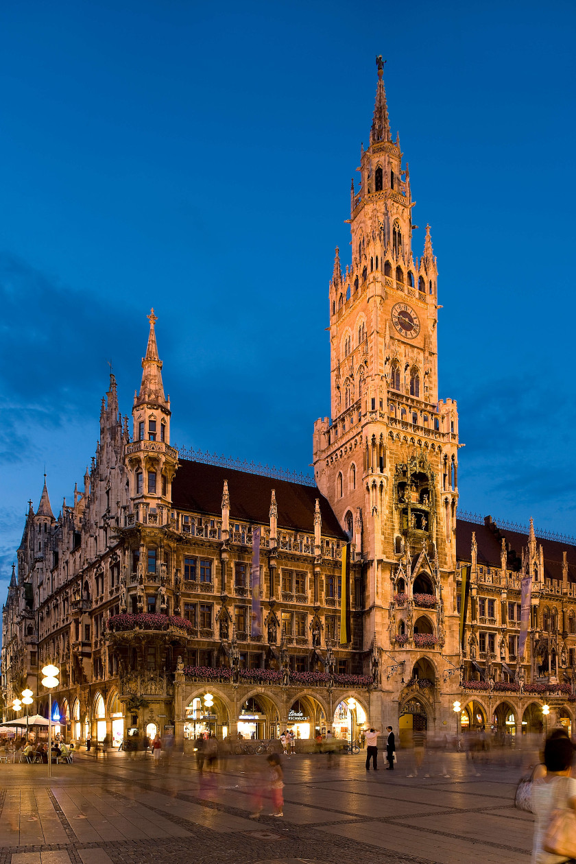 Auf diesem Foto sieht man einen Blick vom Marienplatz auf das Neue Rathaus am Abend. Der Himmel hinter dem Gebäude ist dunkel. Ein Turm in der Mitte der Fassade des Gebäudes ragt in den Himmel. Die Lichter in den Bögen des Gebäudes leuchten hell. Der Platz vor dem Gebäude ist dunkel und vereinzelt sind Personen zu erkennen.