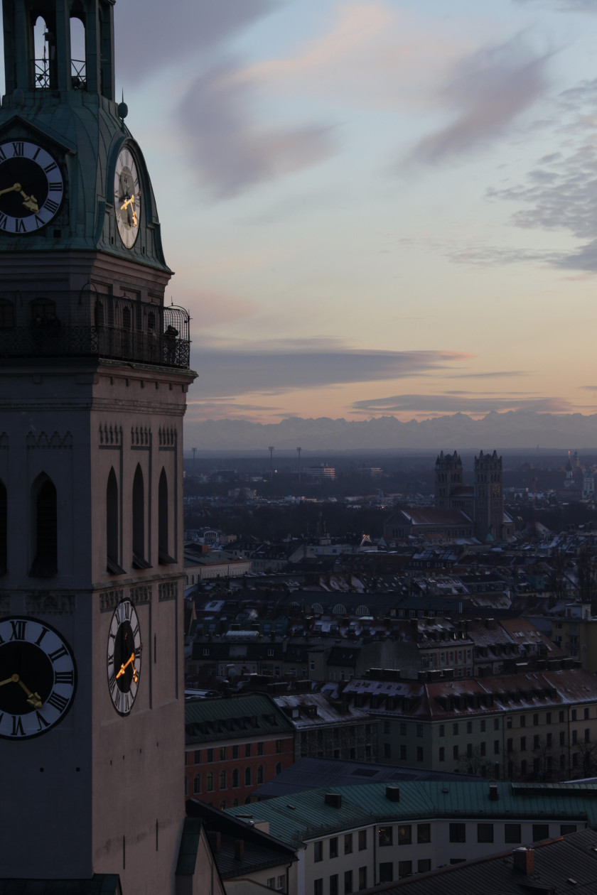 Auf diesem Foto sieht man links im Bild einen Teil des Turms der Sankt Peter Kirche in Nahaufnahme. Das Foto wurde auf Höhe der Aussichtsplattform unter der Turmspitze aufgenommen. Rechts im Bild blickt man von oben aus Vogelperspektive auf die Stadt. Es ist bereits sehr dunkel. Im Hintergrund sieht man in weiter Ferne die Alpen.