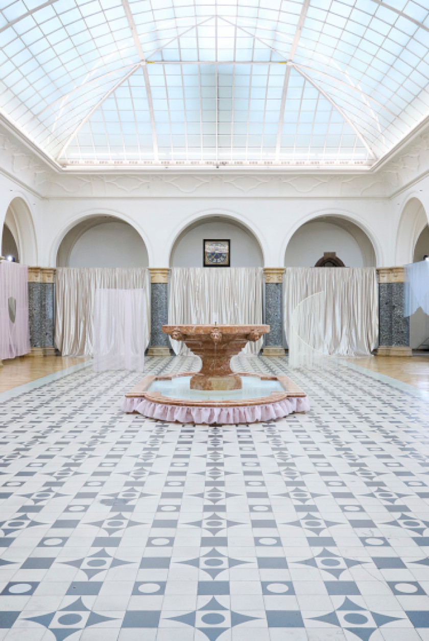 Auf diesem Foto sieht man einen Blick in einen Saal. Die Decke besteht aus einer verglasten Kuppel. In der Mitte des Raumes steht ein Springbrunnen mit Wasser. Der Boden ist aus Fliesen und bildet ein Muster. An den Wänden sind Bögen aus Stein zu sehen. Die Bögen sind auf Säulen. Vor den Bögen hängen Gardinen. Durch die verglaste Kuppel ist der Raum sehr hell.