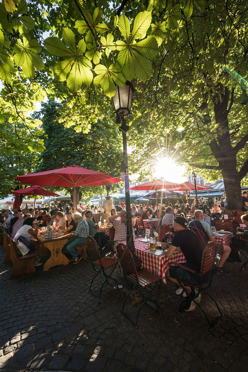 Auf diesem Foto sieht man einen Blick auf den Biergarten des Viktualienmarkts. Viele Menschen sitzen an den Tischen. Über den Tischen sind rote Sonnenschirme. Oben im Bild sind die grünen Blätter von Kastanienbäumen zu sehen und die Sonne scheint.