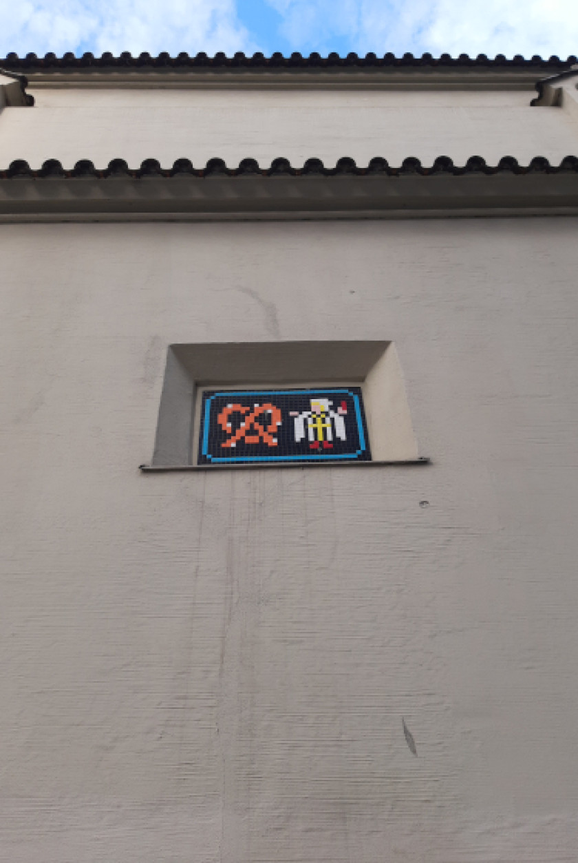 Auf diesem Foto sieht man die Draufsicht und Nahaufnahme einer Mauer. In der Mauer mittig im Bild ist ein Mosaik zu erkennen, das an eine Kachel erinnert. In dieser Kachel sind links eine Breze und rechts daneben das Münchner Kindl aus Mosaik zu erkennen. Das Foto wurde so aufgenommen, als würde man die Mauer hochschauen. Ganz oben im Bild ist noch ein Teil des blauen Himmels zu sehen.