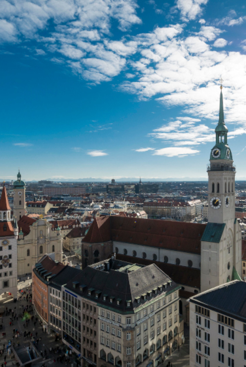 Auf diesem Bild sieht man einen Blick von oben auf München aus der Vogelperspektive. Präsent rechts im Bild ist die Kirche Sankt Peter zu sehen mit dem hohen Turm, der über die umstehenden Dächer herausragt. Der Himmel ist überwiegend blau mit Wolken. Ganz entfernt im Hintergrund sind die Alpen zu erkennen.