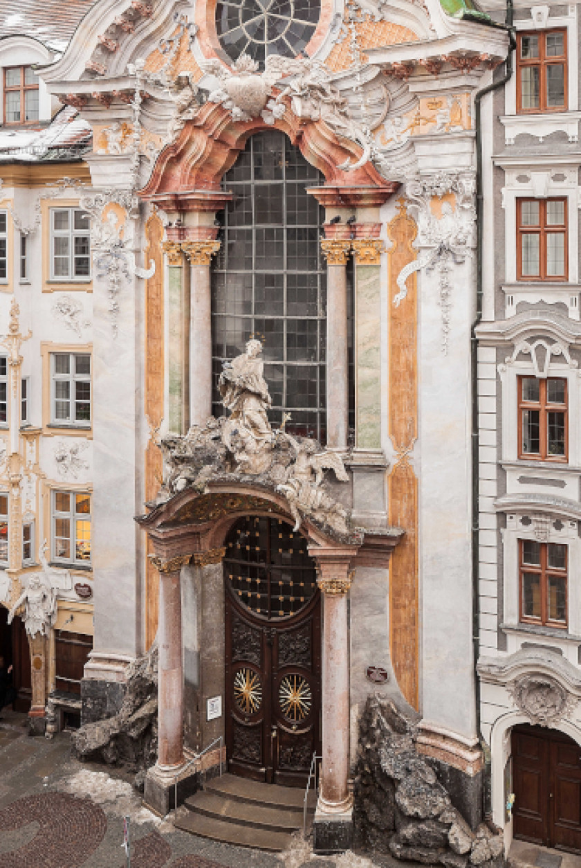 Auf diesem Bild sieht man einen Blick von außen auf eine Kirche. Die Kirche reiht sich in die Häuserfassade der Straße ein. Das Bild wurde von oben geschossen. Unten erkennt man die große Eingangstür der Kirche mit großen Säulen rechts und links. Darüber ist eine große Figur aus Stein und dahinter sind Fenster und Säulen. Die Fassade ist weiß und orange und mit viel Stuck.