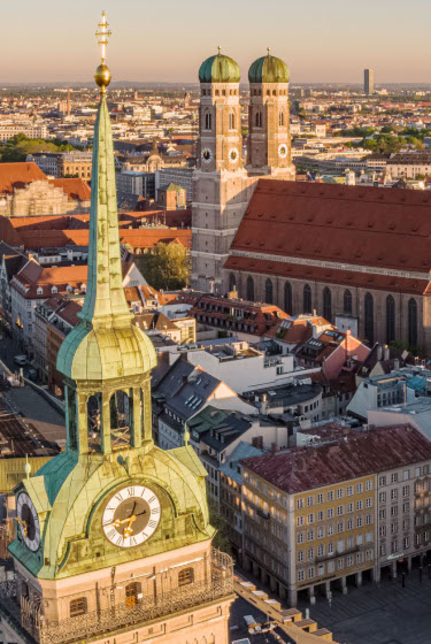 Auf diesem Foto sieht man die grüne Spitze eines Turms im Vordergrund links im Bild von oben fotografiert. Im Hintergrund sieht man die Dächer und Häuser der Stadt. Prominent rechts im Hintergrund des Bildes ist eine große Kirche mit zwei Türmen zu erkennen. Der Blick reicht weit bis in den Horizont.