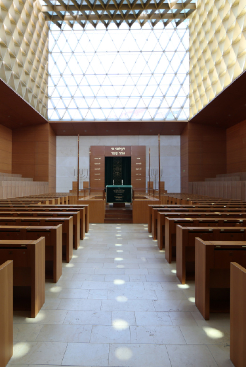 Auf diesem Bild sieht man eine Innenansicht einer Synagoge. Links und rechts im Bild sind Holzbänke zu erkennen. Dazwischen ist ein Gang, der zum Ende der Synagoge führt.