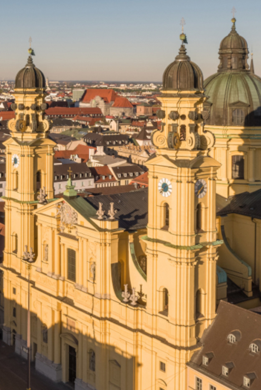 Auf diesem Foto ist die Theatinerkirche in der Vogelperspektive, von schräg oben aufgenommen, zu erkennen. Der größte Teil der Kirche wird von der Sonne angestrahlt. Unten ist ein kleiner Teil im Schatten. Links und rechts neben dem Zentrum der Außenfassade mit Eingang ragen jeweils ein Turm in die Höhe. Die Kirche ist gelb. Dahinter ist eine Kuppel zu erkennen. Im Hintergrund sind Dächer und viele Häuser von oben bis weit in die Ferne zu sehen.