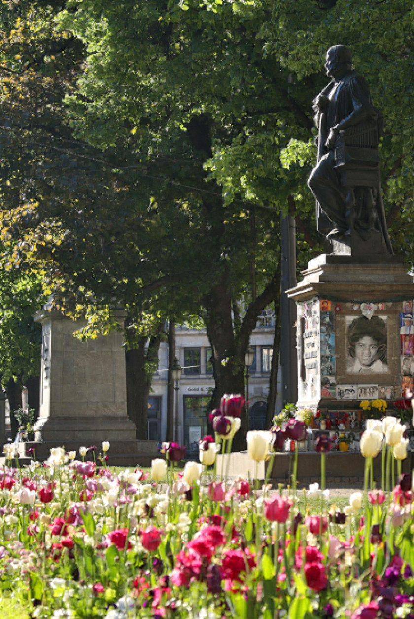 Rechts im Bild ist eine Statue auf einem Sockel aus Stein zu sehen. Die Statue ist von der Seite zu sehen und man erkennt eine männliche Person in der Skulptur. Auf dem Sockel sind außen Fotos von Michael Jackson zu erkennen und auf den Stufen des Sockels sind Blumen und weitere Fotos. Links im Hintergrund des Bildes sind ein weiterer Sockel aus Stein einer Statue zu erkennen sowie Bäume. Im Vordergrund und unten im Bild sind viele blühende Blumen zu sehen.