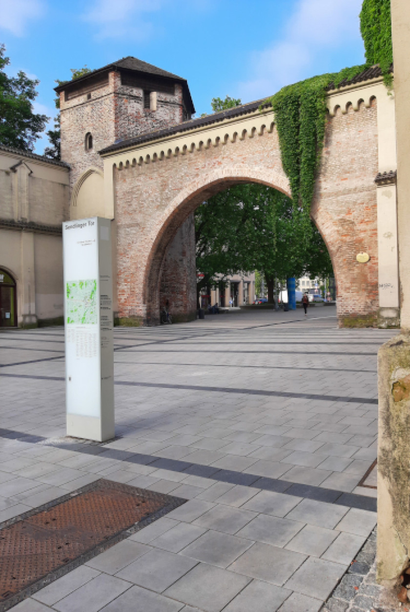 Auf diesem Foto sieht man einen Blick leicht schräg auf das Sendlinger Tor. Links im Bild ist ein Turm zu sehen. Daneben ist ein großer Torbogen. Rechts über dem Torbogen hängt grüner Efeu runter. Auf dem Platz vor dem Tor sieht man eine Stele. Der Platz vor dem Tor ist im Bild sehr präsent und besteht aus grauem Teer. Das Tor ist aus Backsteinziegeln.