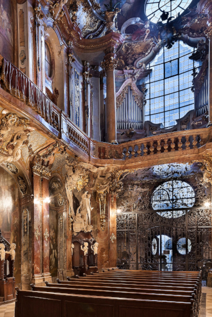 Auf diesem Foto sieht man einen Blick in den Innenraum der Kirche. Man sieht von vorne auf die Bänke aus Holz im Zentrum. Hinter den Bänken sieht man den Eingang der Kirche. Der Innenraum ist verziert mit Stuck und Dekorationselementen. Über dem Eingang ist eine Empore und darüber sind große Fenster.