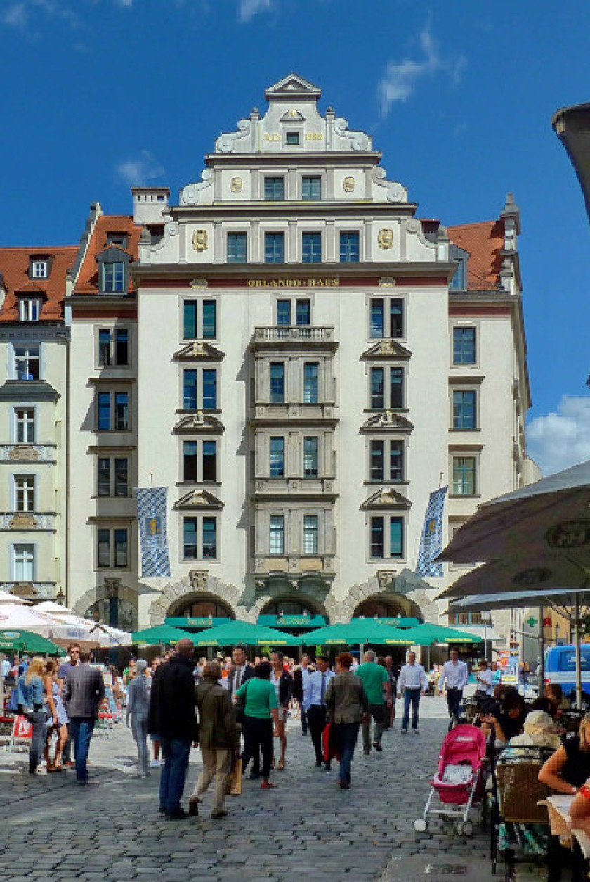 Auf diesem Foto blickt man auf das Orlando-Haus am Platzl. Das Haus ist grün und die Fassade besitzt viele Fenster. Vor dem Haus ist ein belebter Platz mit Stühlen, Tischen, Sonnenschirmen und vielen Personen zu sehen.