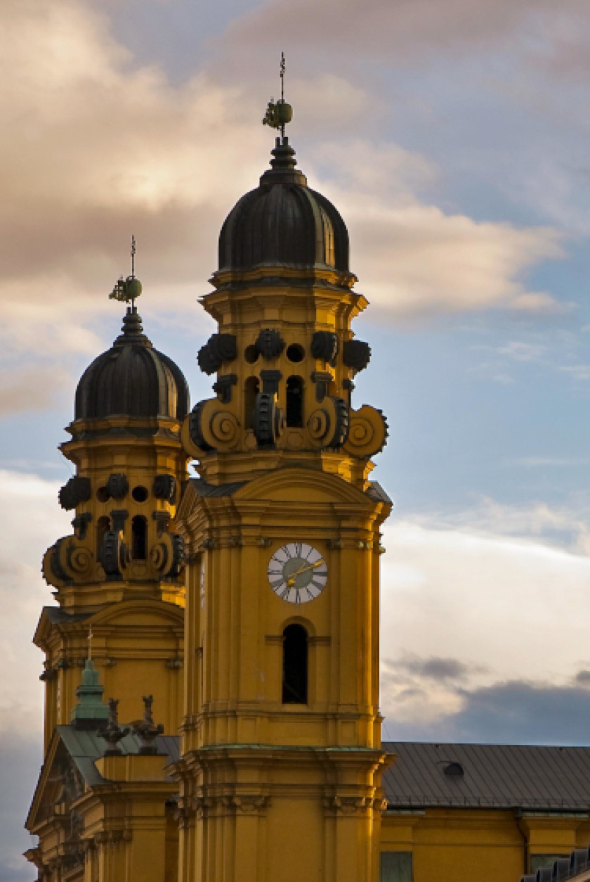 Auf diesem Foto sind zwei Türme im Zentrum des Bildes. Die Türme sind gelb und sind links und rechts an einer Kirche. Das Foto wurde seitlich aufgenommen und man erkennt eine Uhr auf einem Turm. Hinter den Türmen ist der Himmel mit Wolken zu erkennen.