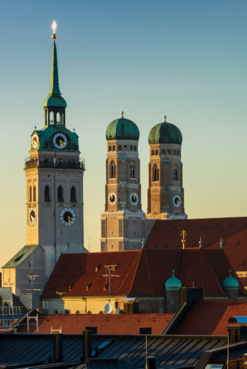 Auf diesem Foto sieht man einen Blick auf die Dächer Münchens im Abendlicht. Zentral im Bild sind Kirchtürme. Links im Bild ist der Kirchturm der Sankt Peter Kirche zu erkennen. Rechts im Bild, schräg dahinter, sind die beiden Türme der Frauenkirche zu sehen. Davor sind die roten Dächer von verschiedenen Häusern auf verschiedenen Höhen zu sehen.