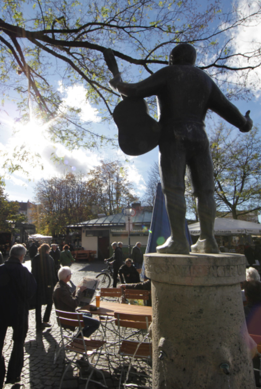 Auf diesem Foto sieht man eine Skulptur auf einem Sockel aus Stein von hinten. Die Skulptur zeigt eine Person mit ausgebreiteten Armen und einer Gitarre in der Hand. Sie befindet sich rechts im Bild. Links im Bild ist ein Blick auf den Viktualienmarkt mit Tischen und Stühlen, Ständen und Personen zu sehen.