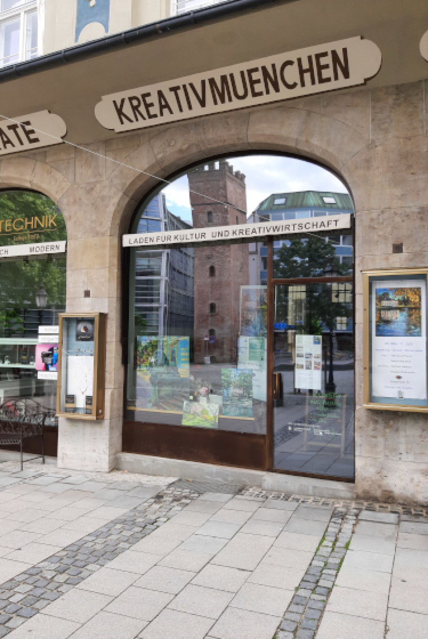Auf diesem Foto sieht man den Kreativ München Pop-up-Store von außen. Über dem Eingang steht in großem Schriftzug: „KREATIVMUENCHEN“. Darunter ist der Eingang in Form eines Bogens. In der Tür spiegelt sich ein Turm. Über der Eingangstür steht noch der Schriftzug „Laden für Kultur und Kreativwirtschaft“. Im Schaufenster, das links im Bild neben der Eingangstür ist, sieht man ausgestellte Bilder. Rechts und links neben dem Eingang und dem Schaufenster sind Infokästen an der Wand. Links im Bild ist noch ein kleiner Teil des Nachbargeschäftes zu erkennen. Unten im Bild und somit vor dem Kreativ München Laden ist noch ein Teil des Platzes vor dem Laden zu erkennen.