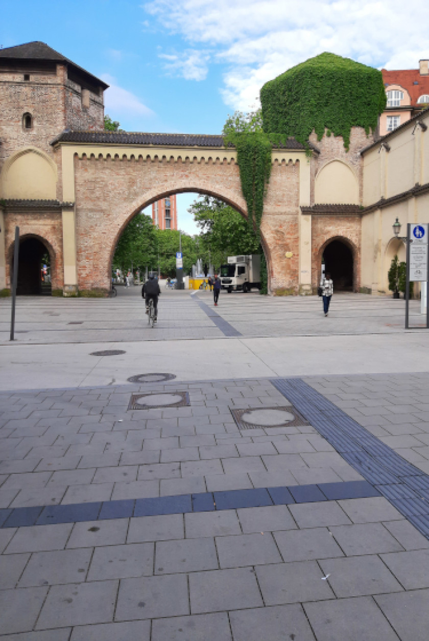 Auf diesem Foto sieht man in der Draufsicht das Sendlinger Tor. Links und rechts an dem Torbogen sind jeweils ein Turm. Auf dem Turm rechts im Bild ist grüner Efeu auf dem Dach, das auf den Torbogen hängt. Das Tor ist aus Backsteinziegeln. Wenn man durch das Tor schaut, sieht man im Hintergrund Bäume, einen Kirchturm und einen LKW. Auf dem Platz vor dem Tor, der einen großen Teil des Bildes einnimmt, sieht man Personen und rechts im Bild ein Schild mit dem Symbol für Fußgänger.