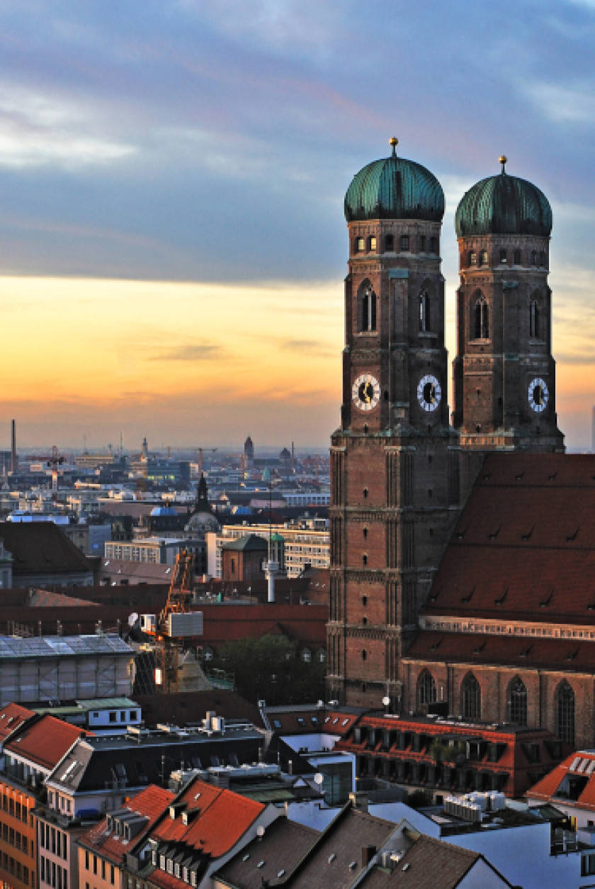 Auf diesem Foto ist ein Teil einer Kirche mit zwei Türmen rechts im Bild von hinten fotografiert. Links im Bild sind Dächer und Gebäude von oben aus der Vogelperspektive zu sehen. Am Himmel kann man die Farben des Sonnenuntergangs oder -aufgangs sehen. Die Kirche und die Dächer der Stadt liegen im Schatten.