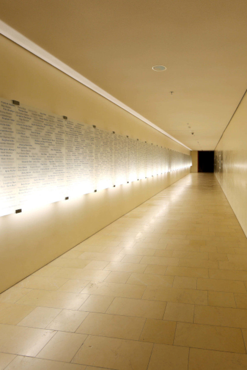 Auf diesem Foto ist ein langer Gang zu sehen. Am Ende des Gangs ist es schwarz. Links an der Wand des Gangs sind beleuchtete Tafeln aus Glas angebracht, auf denen etwas geschrieben ist, was man nicht erkennen kann. Die Wand rechts im Bild neben dem Gang ist leer. Der Gang ist in sehr hellen Farben gehalten und wird nur von der Wandbeleuchtung links beleuchtet.