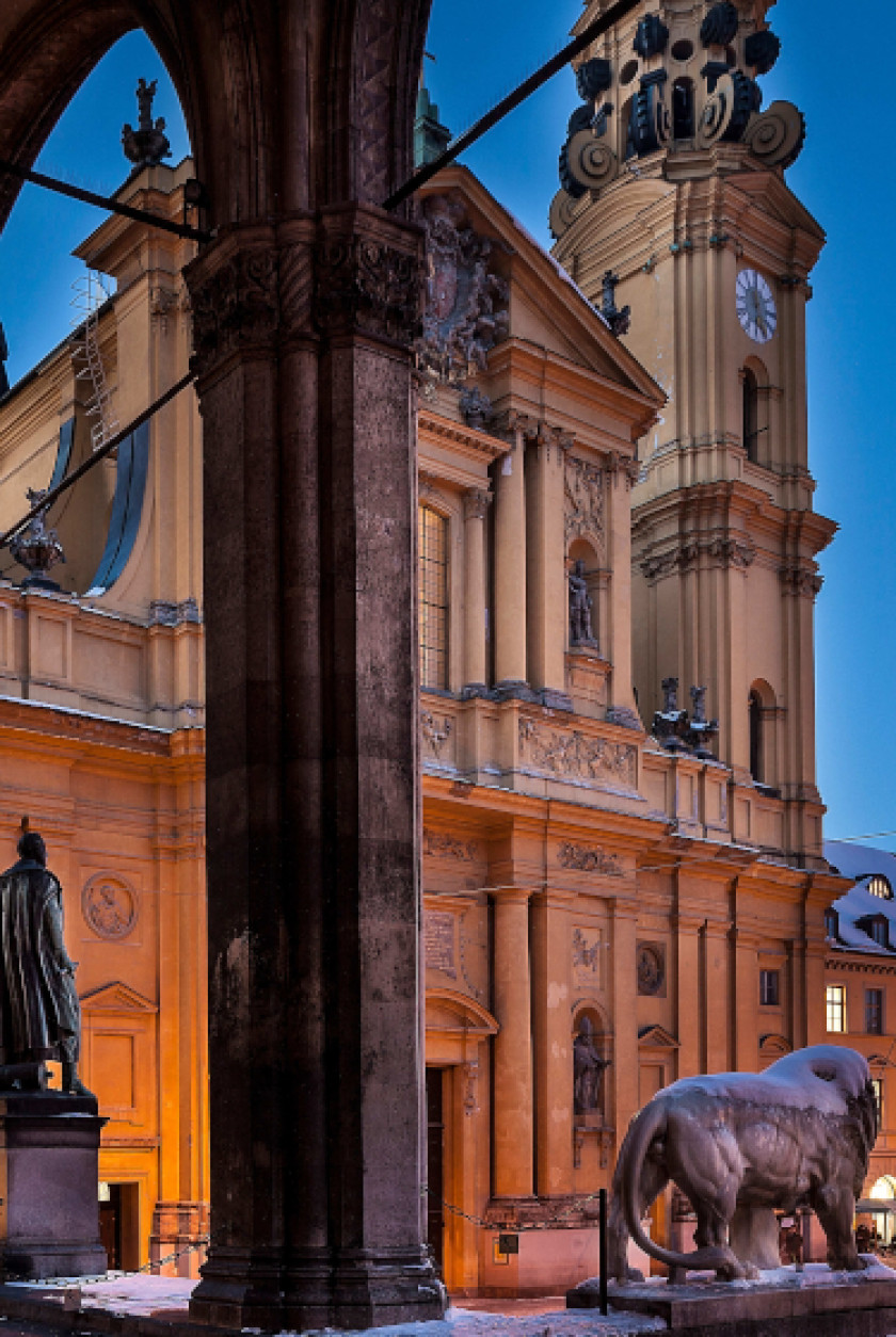 Auf diesem Foto sieht man im Vordergrund ungefähr in der Mitte des Bildes eine Säule, die durchs Bild verläuft. Links davon steht die Statue eines Mannes auf einem Sockel. Rechts davon ist die Statue eines Löwen zu sehen. Die Statuen wurden von hinten fotografiert und sind mit Schnee bedeckt. Im Hintergrund sieht man die gelbe Außenfassade einer Kirche mit einem hohen Turm. Das Foto wurde bei Dämmerung oder vor Sonnenaufgang aufgenommen, da der Himmel im Hintergrund noch dunkel ist.