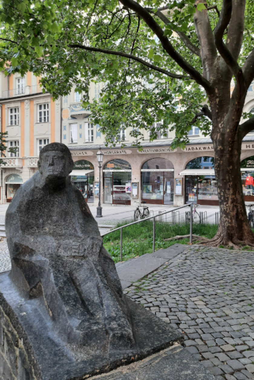 Auf diesem Foto ist eine Skulptur links im Bild im Vordergrund. Die Figur ist menschengroß und sitzt auf einem Stein. Die Figur soll einen Mann darstellen und ist aus Stein oder Marmor. Rechts im Bild dahinter ist ein Baum zu erkennen. Weiter im Hintergrund des Bildes sind verschiedene Geschäfte nebeneinander und eine Treppe zu erkennen.