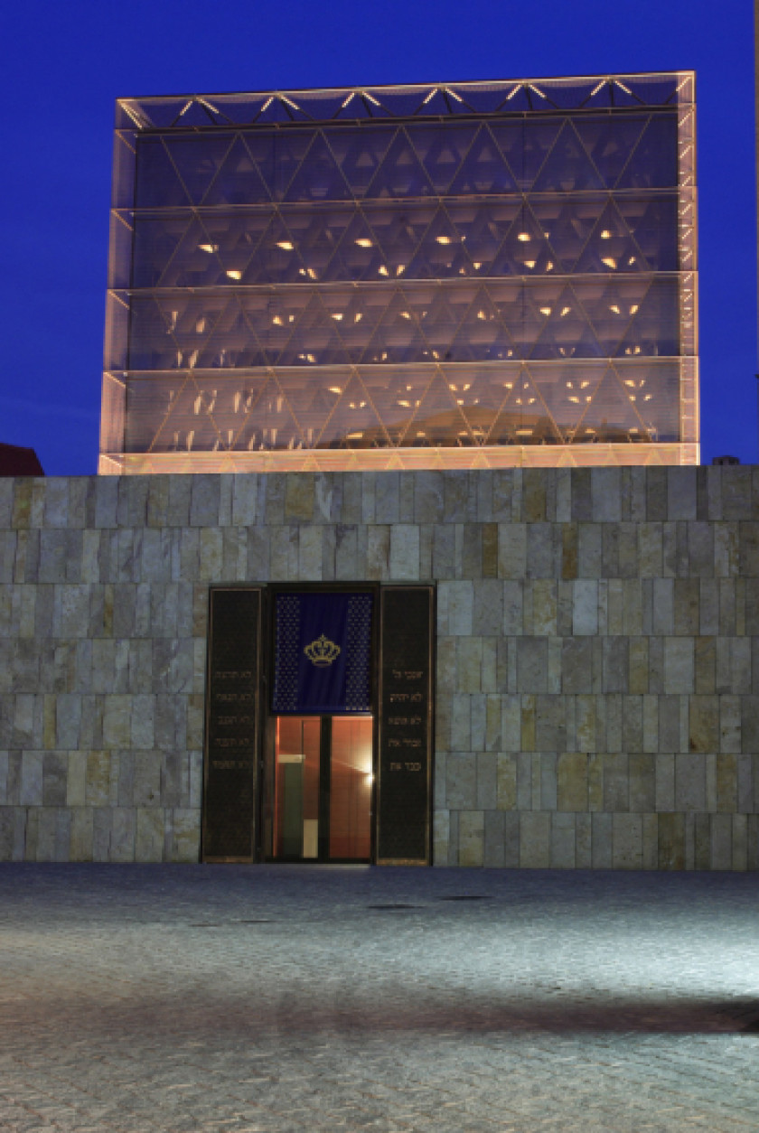 Auf diesem Foto sieht man eine Außenaufnahme der Synagoge bei Nacht. Der Himmel ist dunkel. Die quadratische Kuppel der Synagoge aus Glas ist beleuchtet. Die Außenfassade darunter ist aus Stein. In der Fassade ist die Eingangstür der Synagoge zu sehen. Hinter der Eingangstür aus Glas sieht man Licht. Vor der Synagoge ist unten im Bild noch ein Teil des Vorplatzes zu sehen.