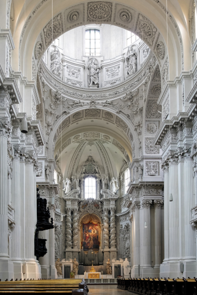 Auf diesem Foto sieht man die Innenansicht einer Kirche. Links und rechts sieht man Sitzreihen von hinten aufgenommen. Dazwischen führt ein Gang entlang zum Altar. Links und rechts außen im Bild neben den Sitzreihen sind weiße Säulen mit Stuck zu sehen. Oben im Bild ist ein weißer Bogen zu sehen, und dahinter ist eine Kuppel zu erkennen, durch die helles Licht scheint. Das Bild hinter dem Altar ist beleuchtet. Der Innenraum ist ganz in Weiß  gehalten.