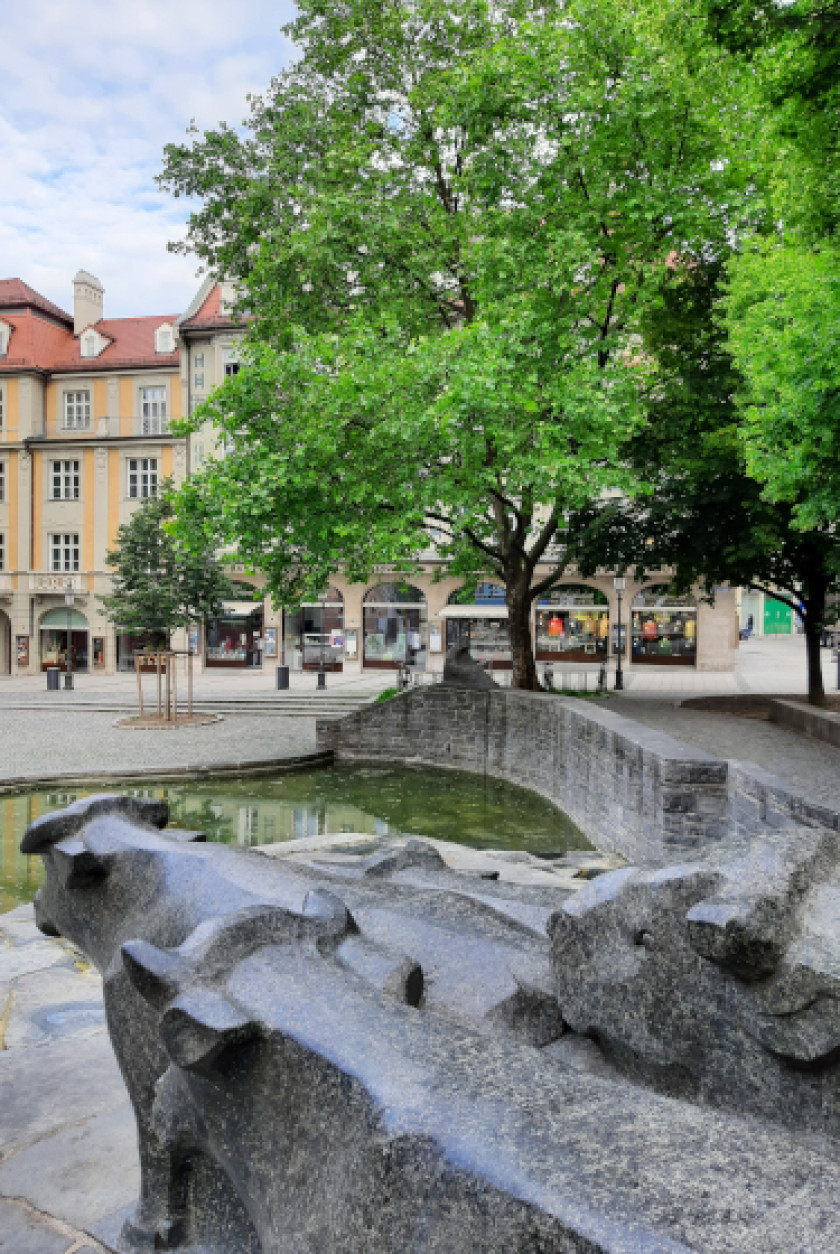 Auf diesem Foto sieht man einen Blick auf den Rindermarkt. Unten im Bild im Vordergrund sieht man Skulpturen von Kühen aus Stein oder Marmor von hinten. Sie schauen in Richtung des Marktes und werden von hinten seitlich fotografiert. Vor den Skulpturen und somit mittig im Bild ist ein kleiner grüner See zu erkennen. Darüber führt rechts im Bild eine Brücke aus Stein. Wenn man über die Brücke geht, gelangt man auf einen Platz. Auf dem Platz sind rechts im Bild zwei große, grüne Laubbäume. Dahinter sind verschiedene Geschäfte nebeneinander zu erkennen. Links im Bild im Hintergrund ist ein gelbes Haus zu sehen.