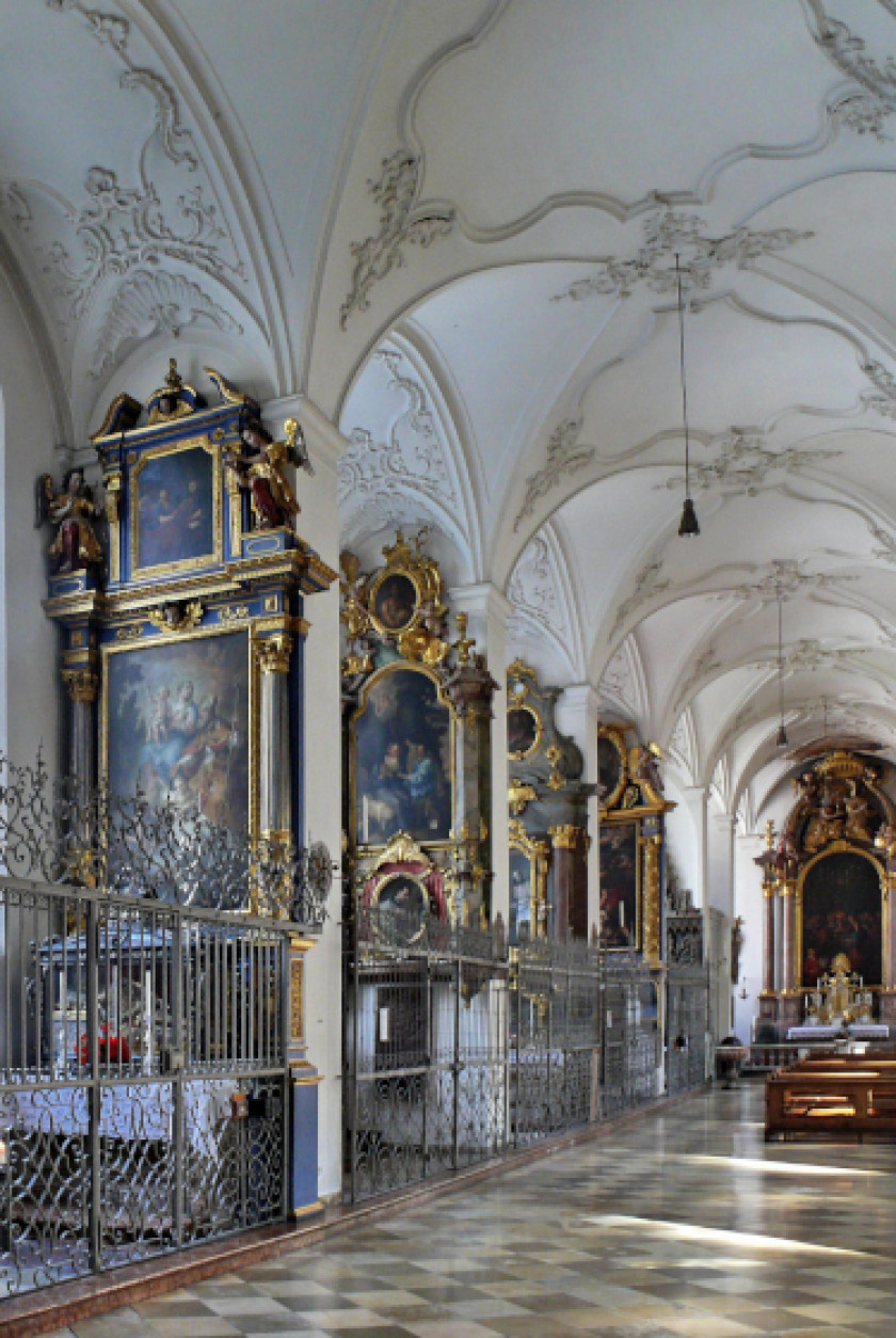 Auf diesem Foto sieht man den Innenraum der Kirche. Links im Bild sind verschiedene kleinere Seitenaltäre nebeneinander zu sehen, die mit einem Gitter abgetrennt sind. Rechts im Bild ist im Hintergrund eine Stuhlreihe zu erkennen, die ebenfalls zu einem Altar ausgerichtet ist. Die Decke ist weiß und stuckverziert.