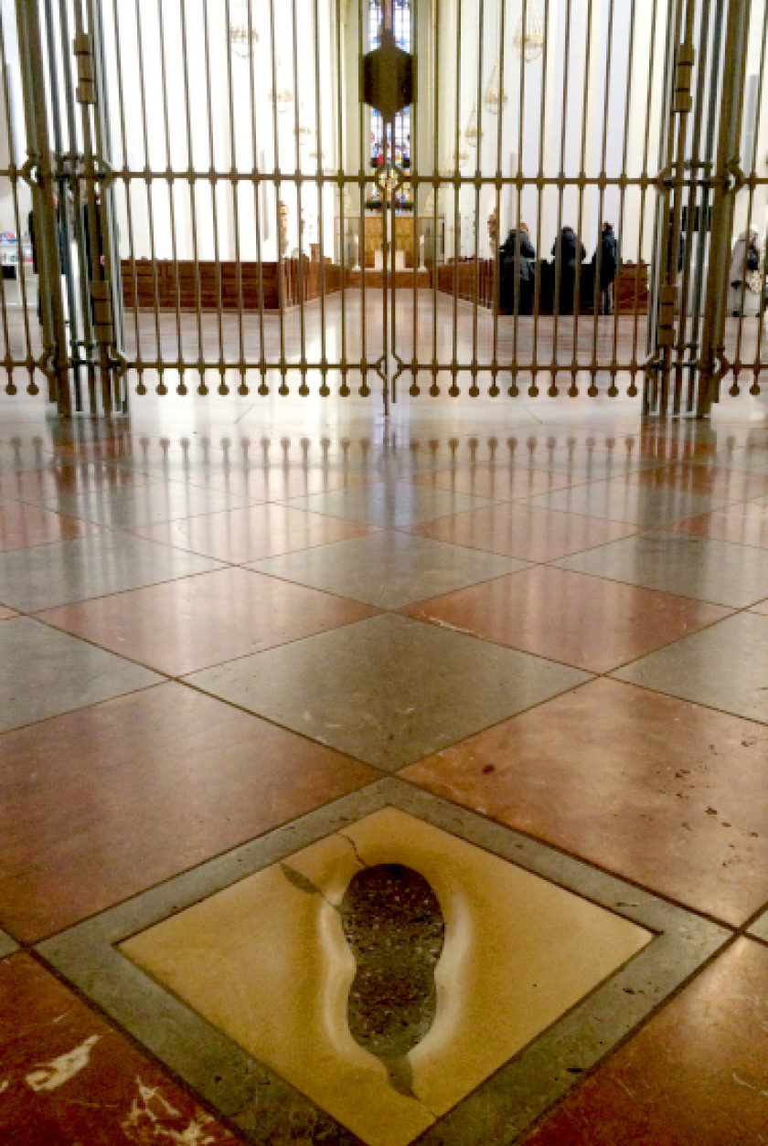Auf diesem Foto sieht man den gefliesten Boden des Innenraums einer Kirche. Im Vordergrund mittig im Bild wurde ein Fußabdruck im Boden fotografiert. Der Fußabdruck ist quadratisch und Gold umrahmt. Im Hintergrund sind Gitterstäbe zu erkennen.