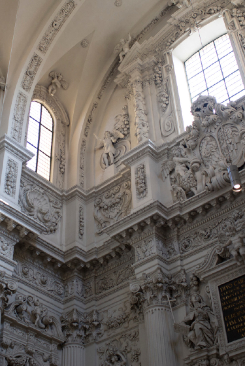 Auf diesem Foto sieht man eine Nahaufnahme eines Teils des Innenraums der Kirche. Man kann die Architektur erkennen: Weißer Stuck und viele herausstehende Verzierungen. Das Bild wurde von unten nach oben geschossen. Oben im Bild sind auch noch Fenster zu sehen.